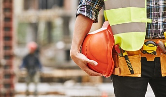 Construction worker. Cropped photo of male professional builder in working uniform with construction tools holding a safety red helmet while standing outdoor of construction site